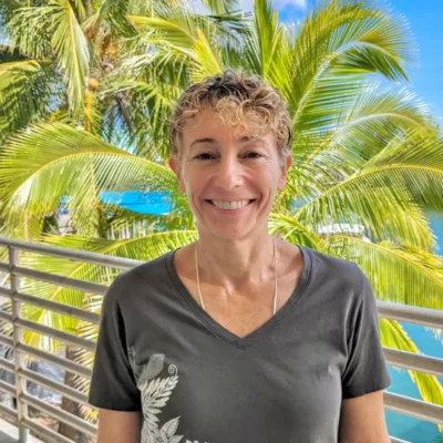 Smiling person with short curly hair stands outside by palm trees and water on a sunny day.
