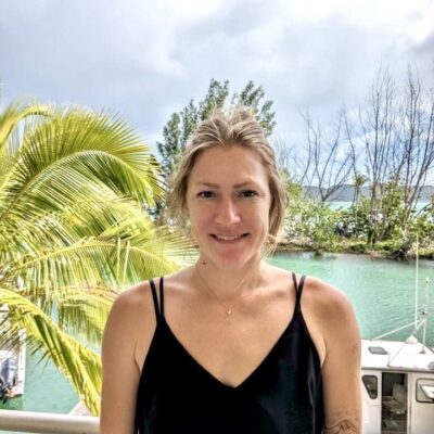 Woman in a black tank top stands by water with palm trees and a boat in the background, smiling at the camera.