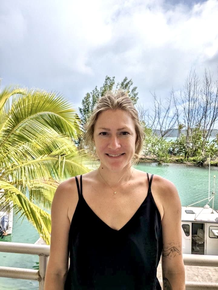 Woman in a black tank top stands by water with palm trees and a boat in the background, smiling at the camera.