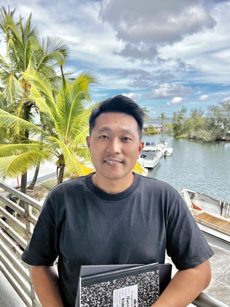 Smiling person in a black shirt holding notebooks by a riverside with palm trees and boats in the background.