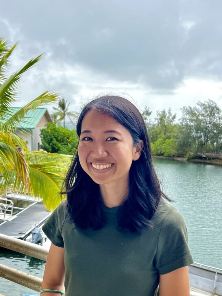 Woman with dark hair smiling by a waterfront, with palm trees and boats in the background on a cloudy day.