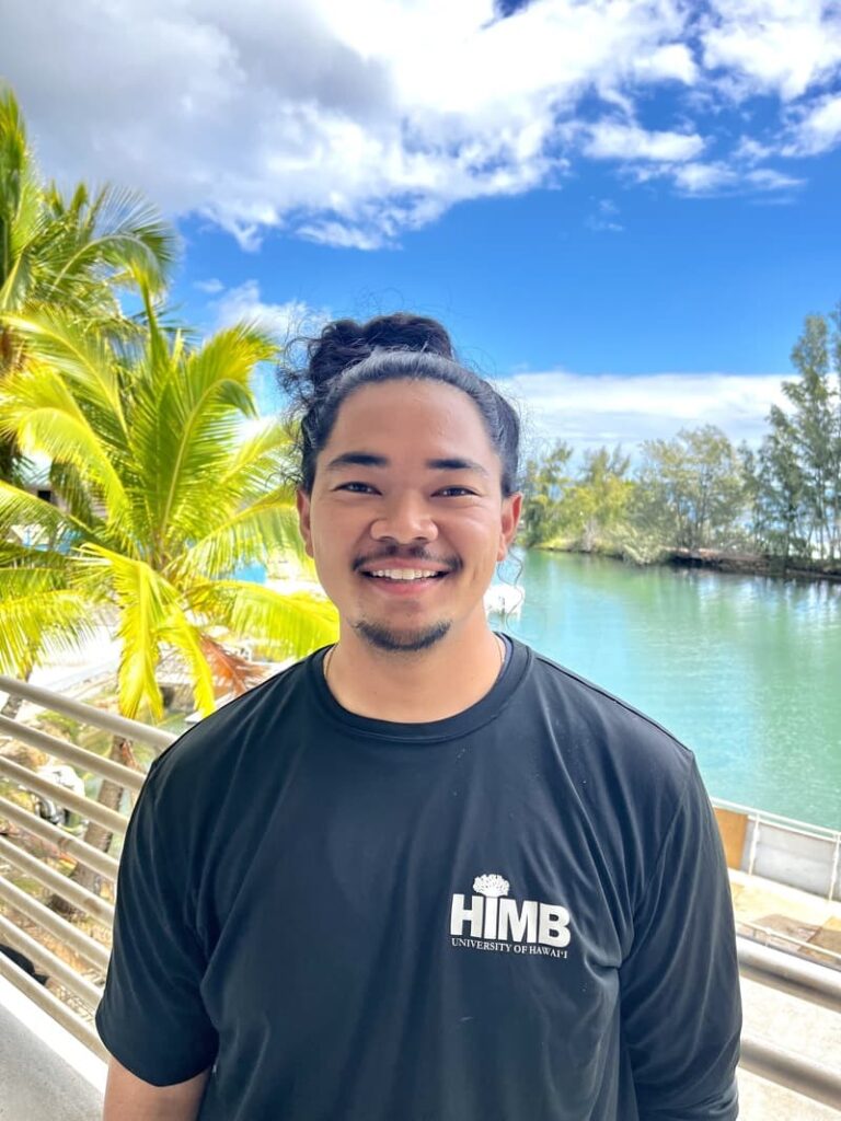 Smiling person in a black HIMB shirt stands by water, palm trees, and blue sky with clouds in the background.