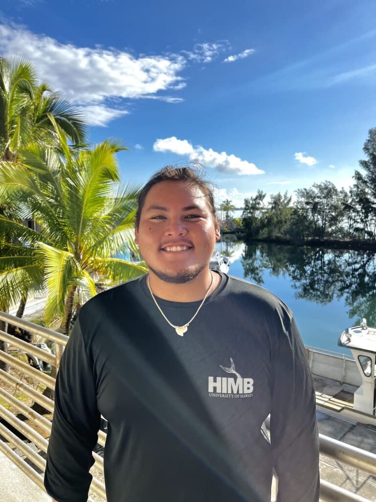 Smiling person in a black HIMB shirt stands by water, palm trees, and blue sky with clouds in the background.