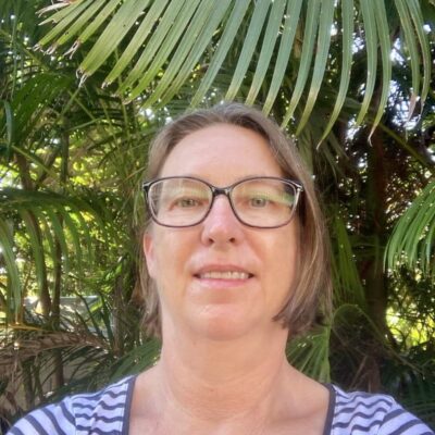 A woman with glasses smiles in front of lush green palm leaves outdoors.