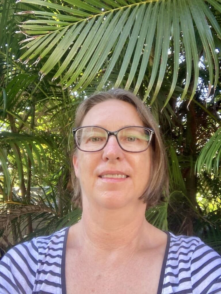A woman with glasses smiles in front of lush green palm leaves outdoors.