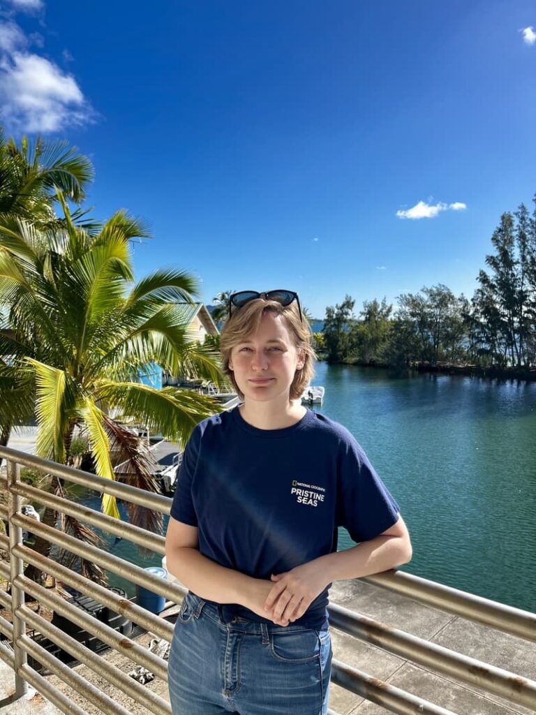 A person stands by a palm tree and water, wearing a navy shirt and sunglasses on their head, under a blue sky.