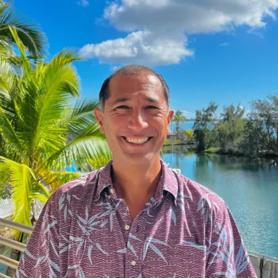 Smiling man in a patterned shirt stands by a tropical lake with palm trees and blue sky in the background.