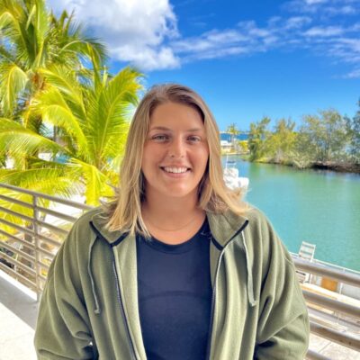 Smiling woman in a green jacket stands on a balcony with palm trees and water in the background.