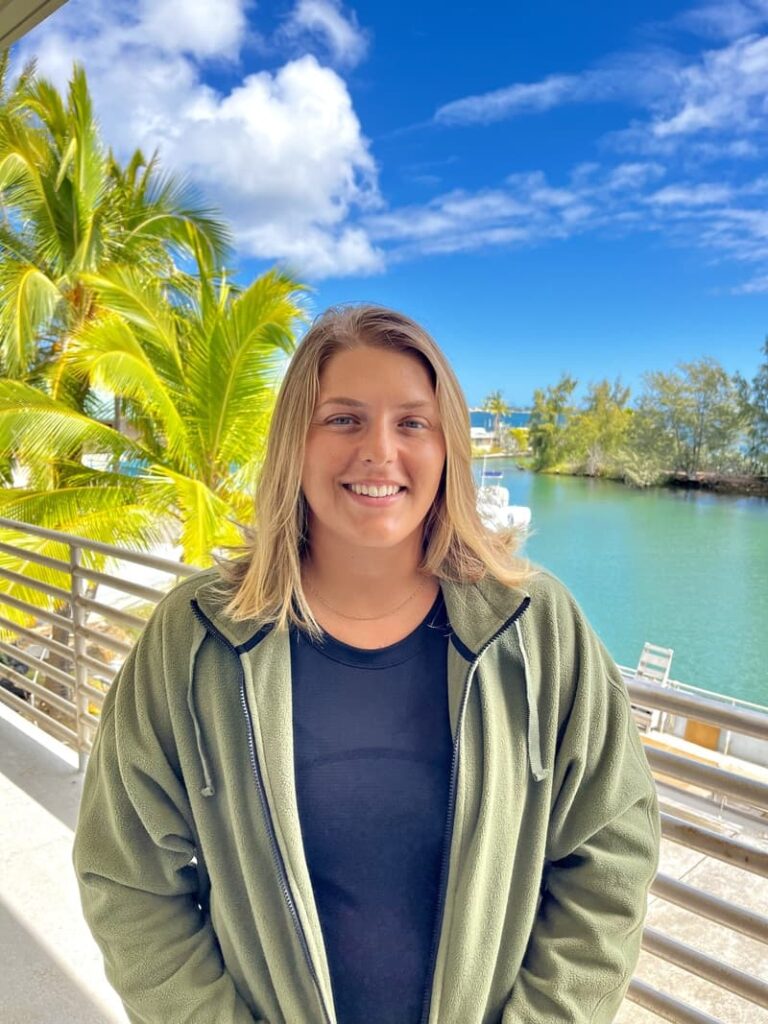 Smiling woman in a green jacket stands on a balcony with palm trees and water in the background.