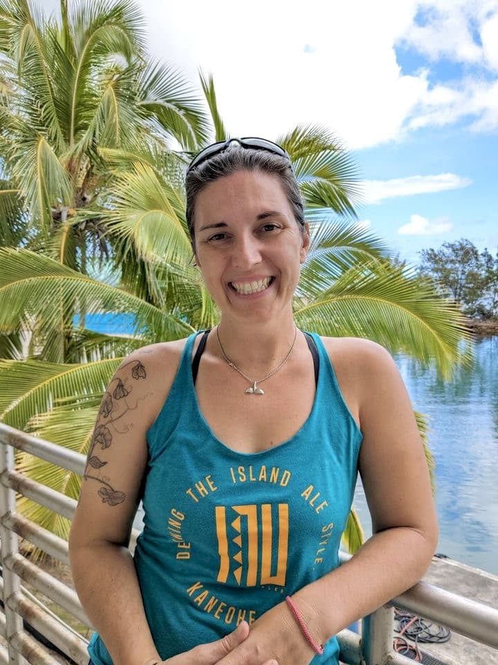 Smiling woman in a teal tank top stands by water with palm trees in the background on a sunny day.