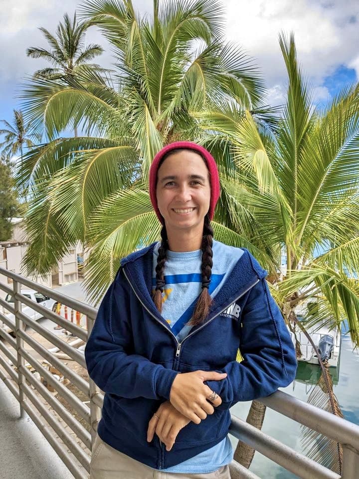 Smiling person in a red beanie and blue jacket stands by a railing with palm trees and water in the background.