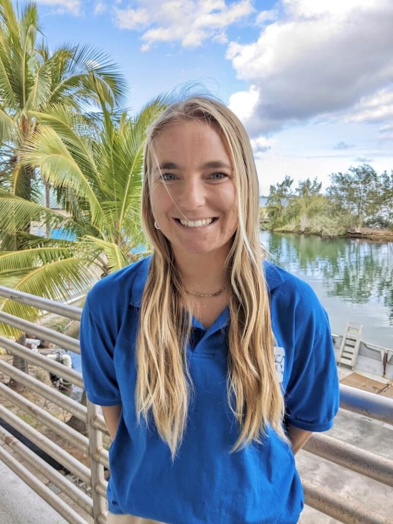 Smiling woman in a blue polo shirt stands by a railing with palm trees and water in the background.