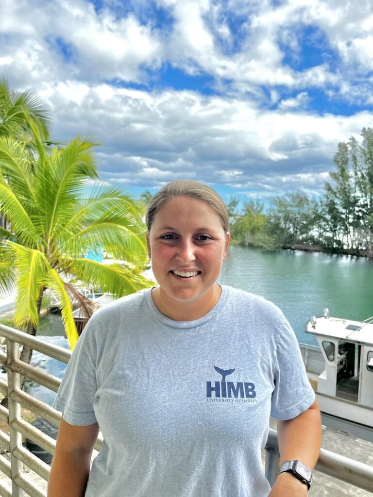 A person in a gray HIMB shirt smiles by water with boats, palm trees, and a cloudy blue sky in the background.