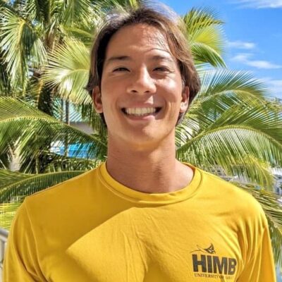 Smiling person in a yellow HIMB shirt stands outdoors with palm trees and blue sky in the background.