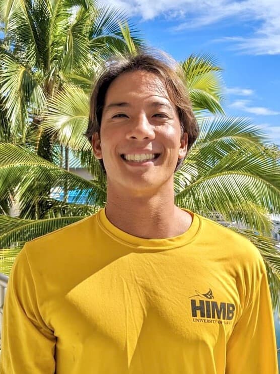Smiling person in a yellow HIMB shirt stands outdoors with palm trees and blue sky in the background.