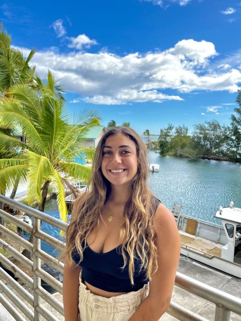 Smiling woman with long hair stands by a railing with water, a boat, palm trees, and blue sky behind her.