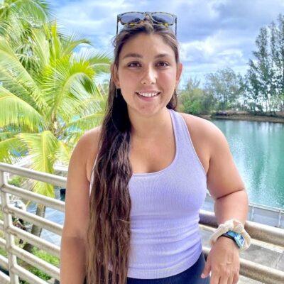 Woman with long brown hair in a tank top stands on a balcony by water and palm trees, smiling at the camera.