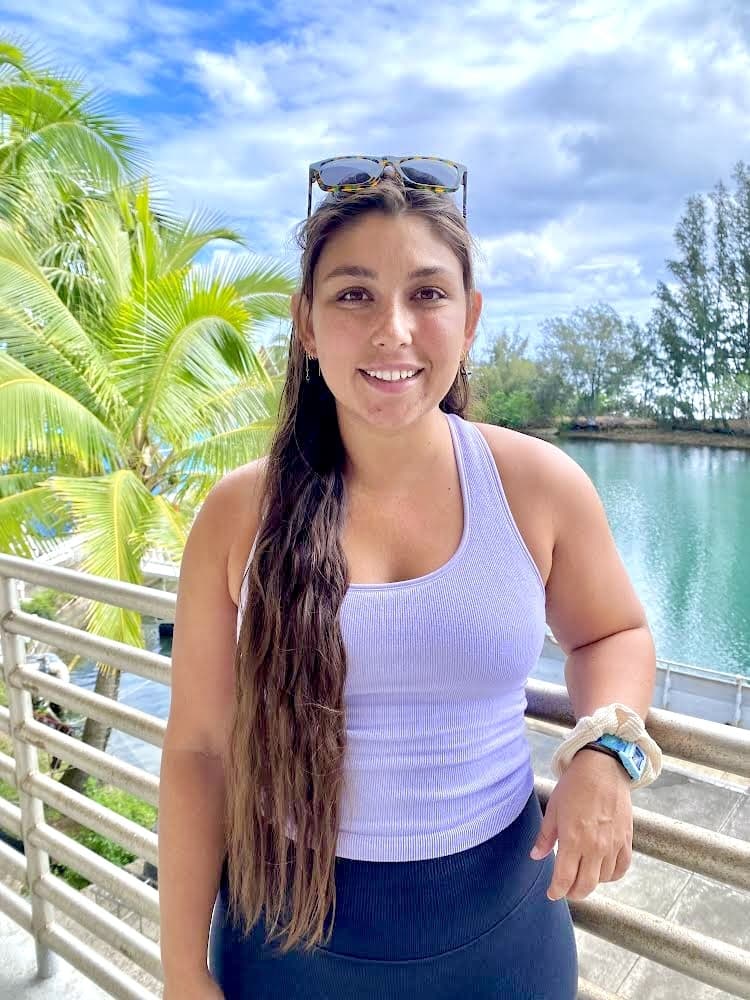 Woman with long brown hair in a tank top stands on a balcony by water and palm trees, smiling at the camera.
