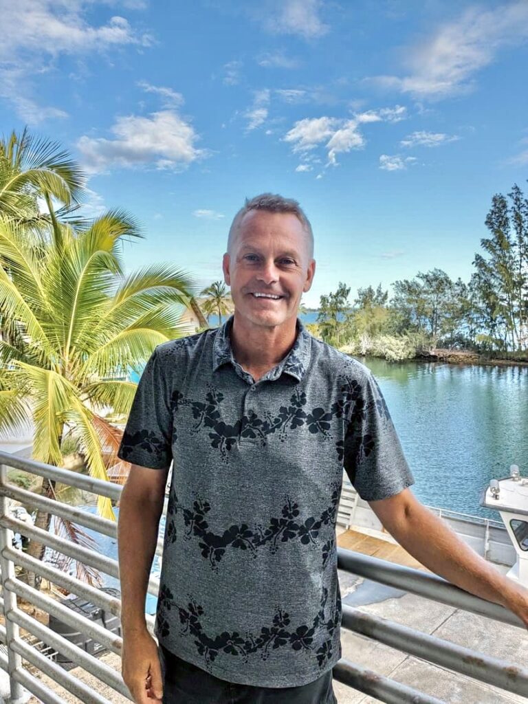 Smiling man in a floral shirt stands by a railing with palm trees and water in the background.