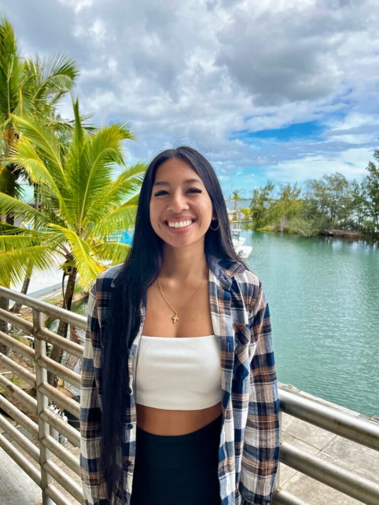 Smiling woman in a plaid shirt stands by a railing with water, palm trees, and cloudy sky in the background.