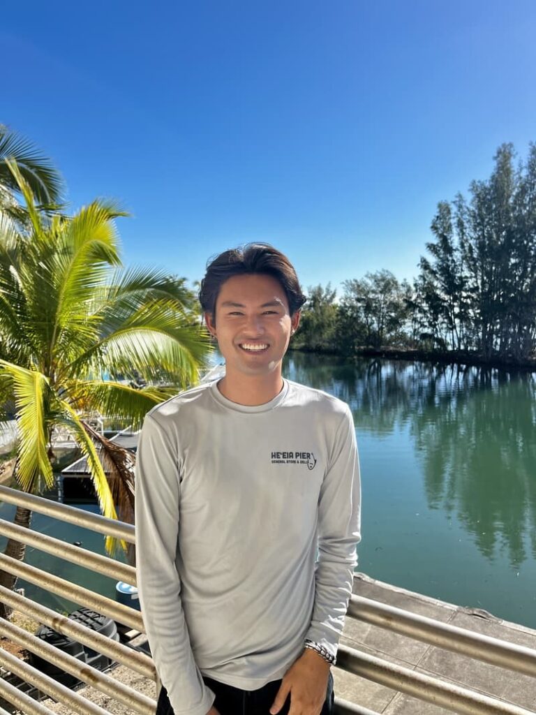 A man in a gray shirt smiles by a railing with water, palm trees, and a clear blue sky in the background.