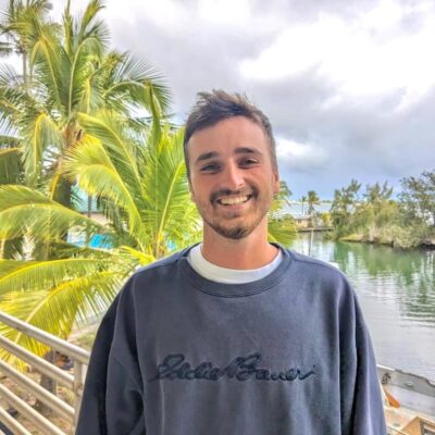 Smiling young man in a navy sweatshirt standing by palm trees and water on a cloudy day.