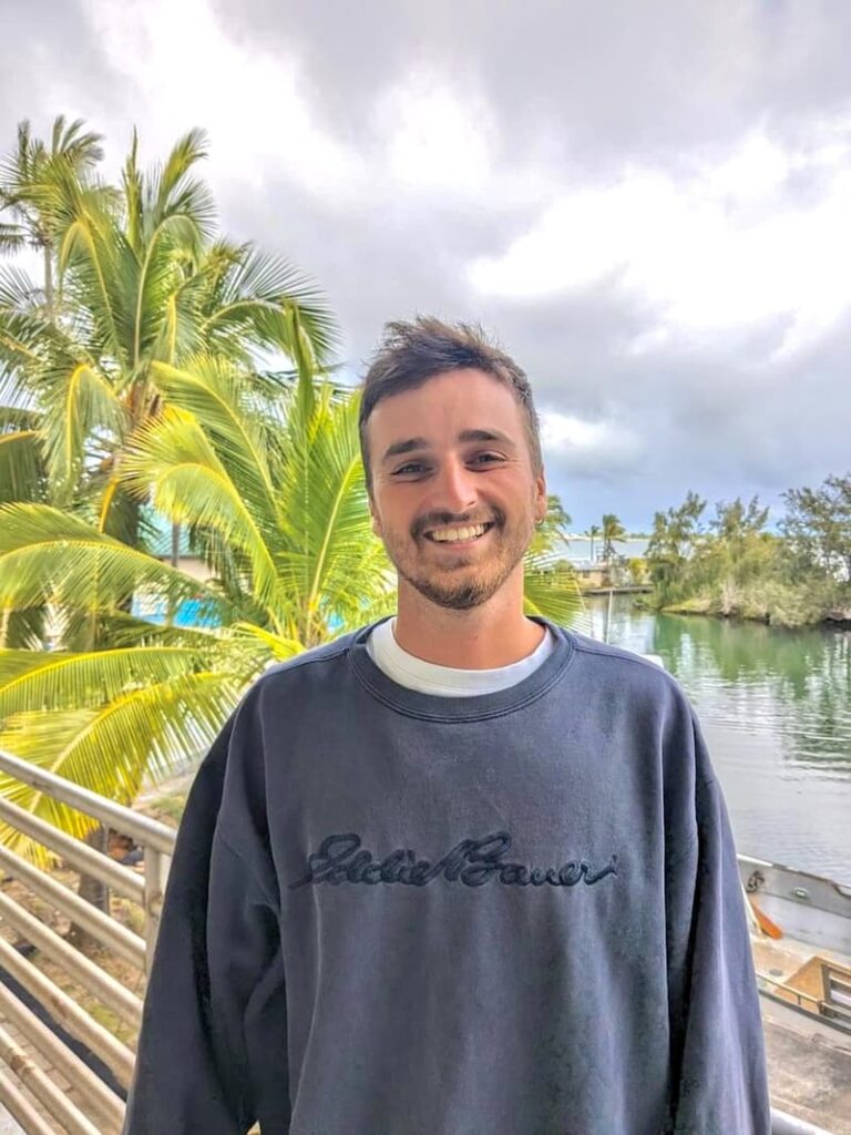 Smiling young man in a navy sweatshirt standing by palm trees and water on a cloudy day.