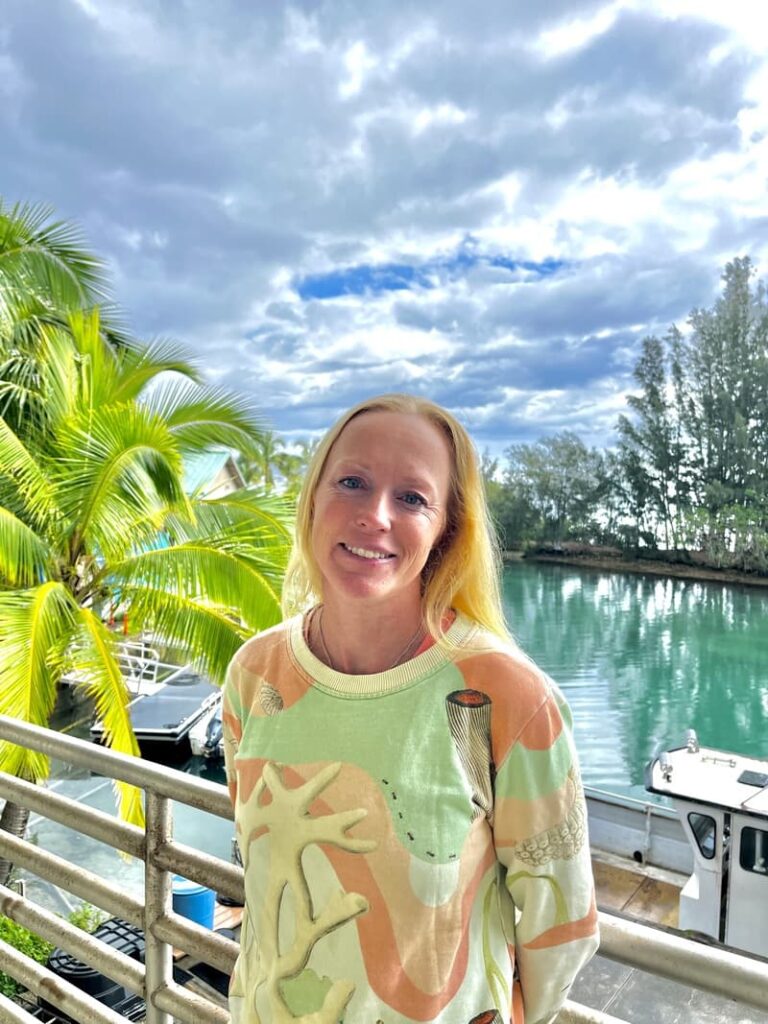 A woman stands by a railing near water with boats, palm trees, and a cloudy sky in the background.