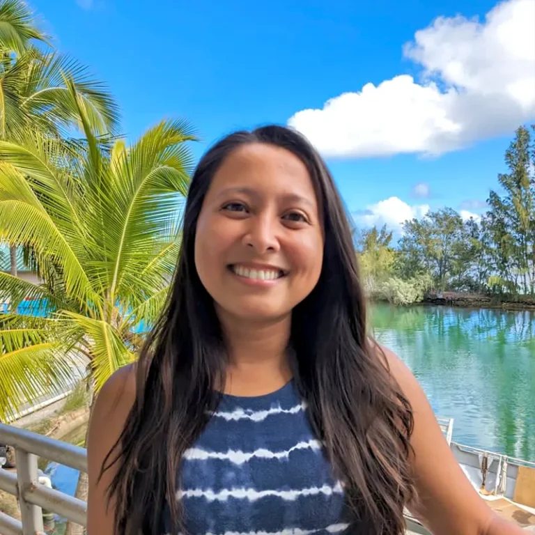 Smiling woman with long hair stands by tropical water, palm trees, and blue sky in the background.