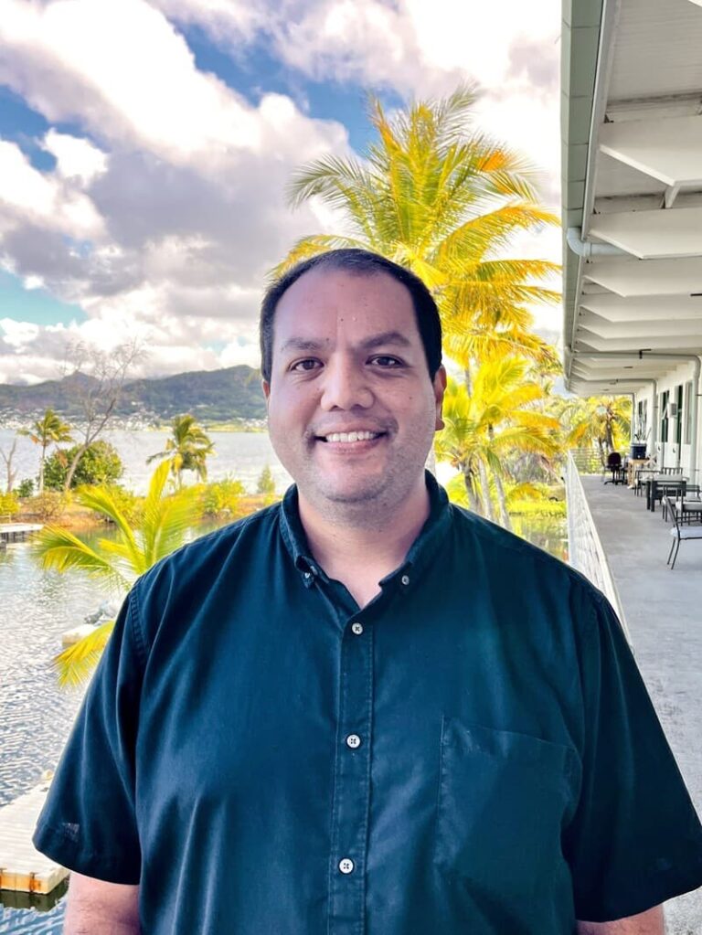 Smiling man in a black shirt stands on a balcony with palm trees and water in the background.