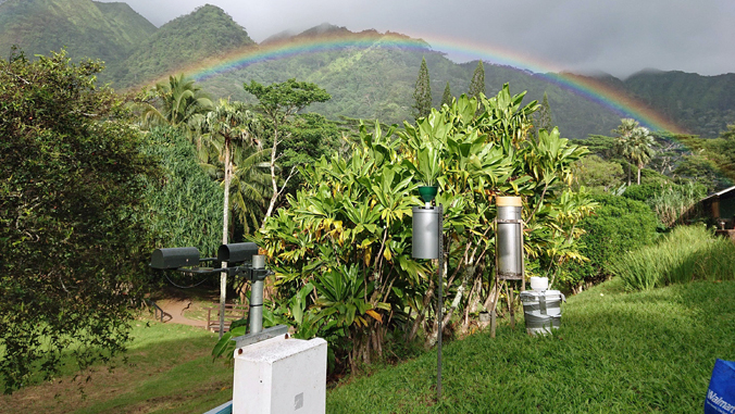Lush green landscape with weather instruments and a rainbow arching over mountains in the background.