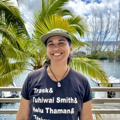 Smiling person wearing a black T-shirt and cap stands outdoors with palm trees and water in the background.