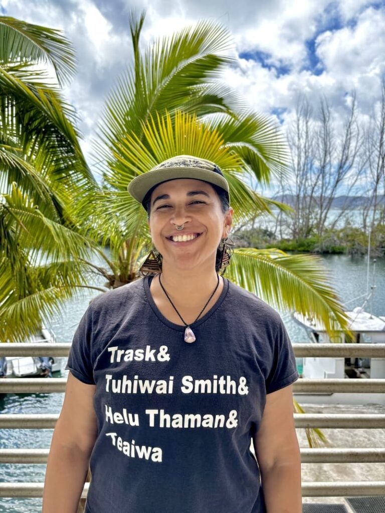 Smiling person wearing a black T-shirt and cap stands outdoors with palm trees and water in the background.
