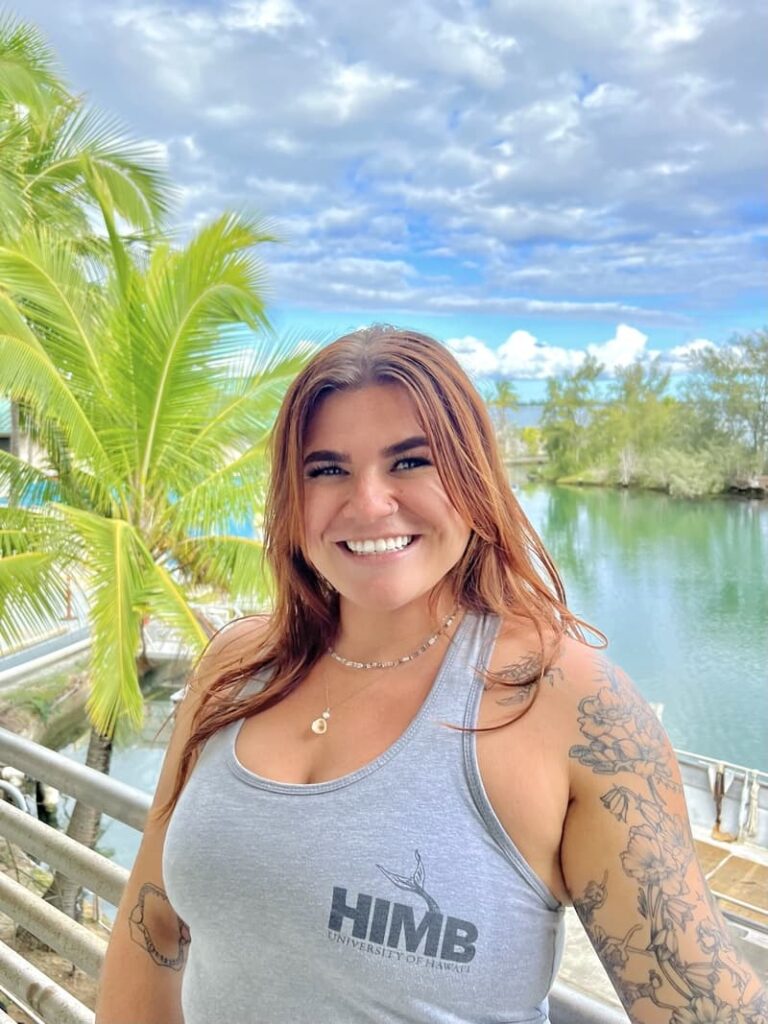 Smiling woman in a gray HIMB tank top stands by water with palm trees and a partly cloudy sky behind her.