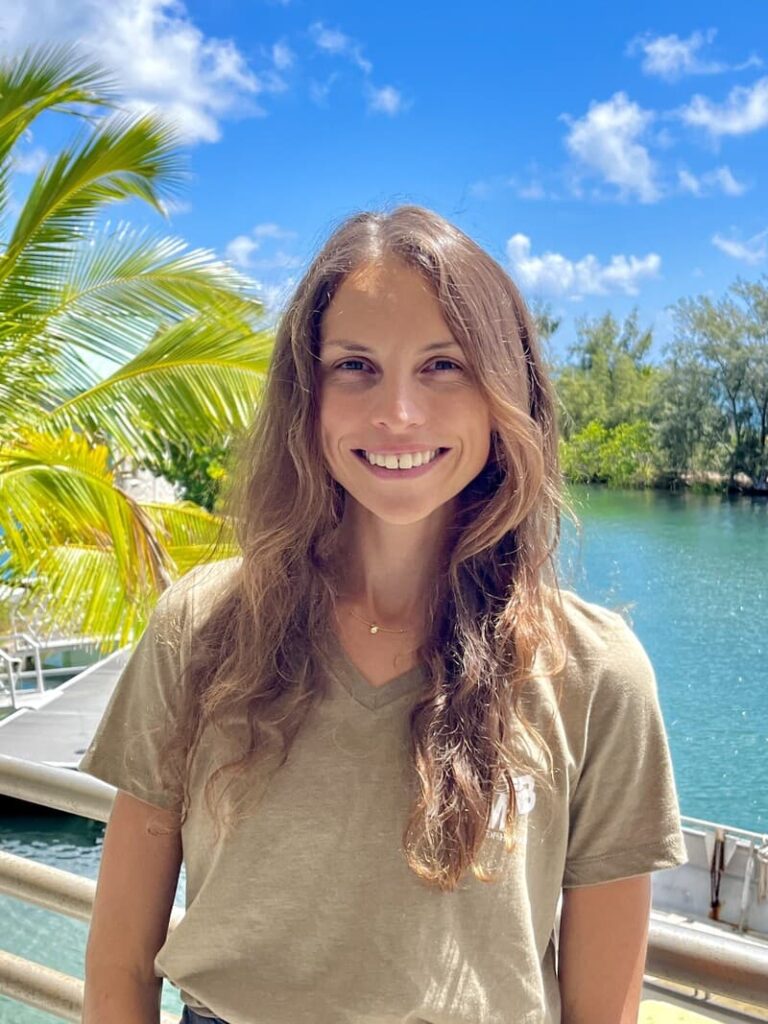 Smiling woman with long hair stands by water, palm trees, and blue sky with clouds in the background.
