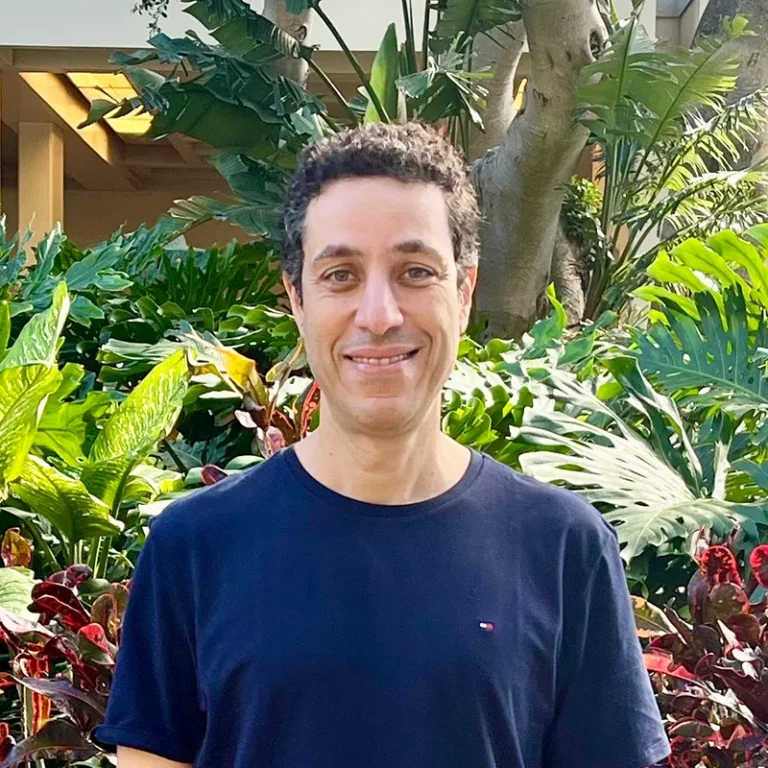 A person in a navy t-shirt smiles while standing in front of lush green tropical plants outdoors.