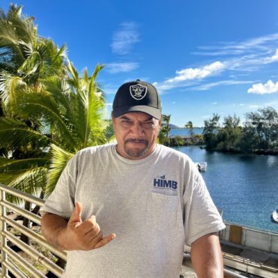 Man in grey HIMB shirt and Raiders cap making shaka sign, standing by water and palm trees under blue sky.
