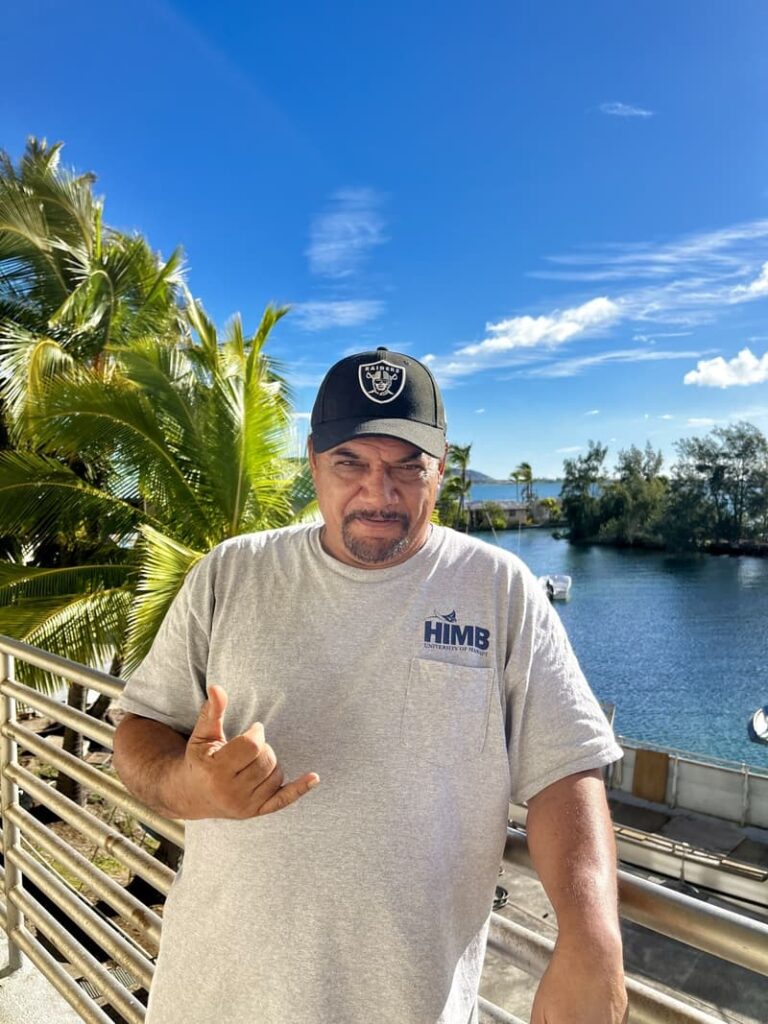 Man in grey HIMB shirt and Raiders cap making shaka sign, standing by water and palm trees under blue sky.