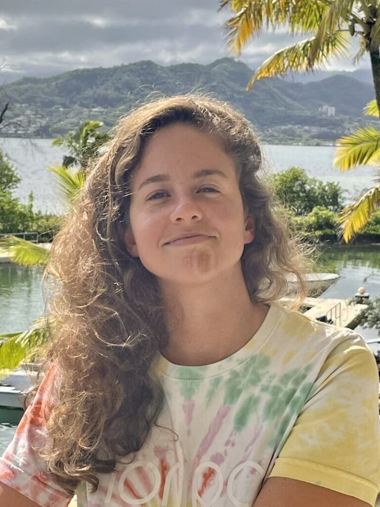 Young woman with curly hair smiles outdoors by a lake, with mountains and palm trees in the background.