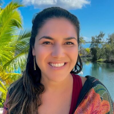 Smiling woman with long dark hair stands outdoors by water, palm trees, and blue sky with clouds.