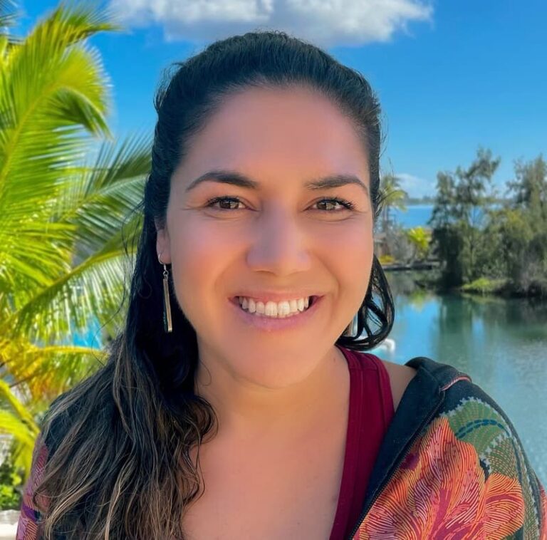 Smiling woman with long dark hair stands outdoors by water, palm trees, and blue sky with clouds.