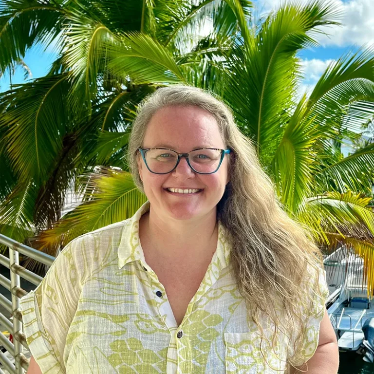 Smiling woman with long hair and glasses stands outdoors in front of palm trees on a sunny day.