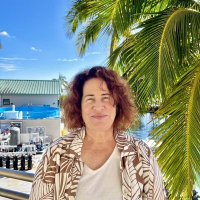 Woman smiling outdoors by palm trees with blue sky, water, and buildings in the background.