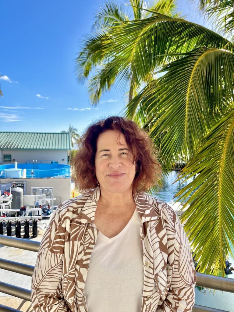 Woman smiling outdoors by palm trees with blue sky, water, and buildings in the background.
