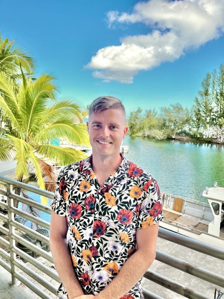 Smiling man in a floral shirt stands by a waterfront with palm trees and a partly cloudy sky.