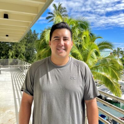 Man in a gray shirt smiling outdoors on a sunny day with palm trees and blue sky in the background.