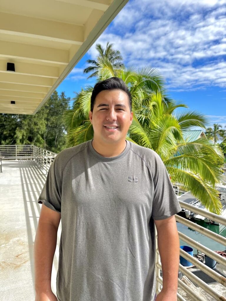 Man in a gray shirt smiling outdoors on a sunny day with palm trees and blue sky in the background.