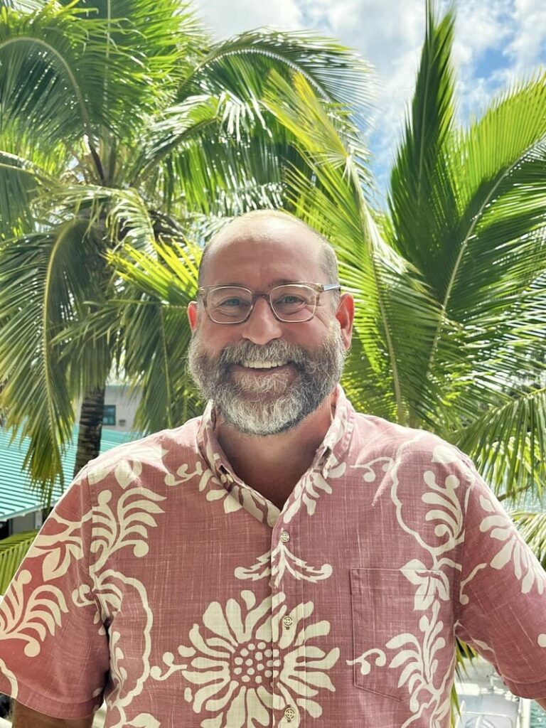 Smiling man with glasses and beard in a pink floral shirt standing in front of palm trees on a sunny day.