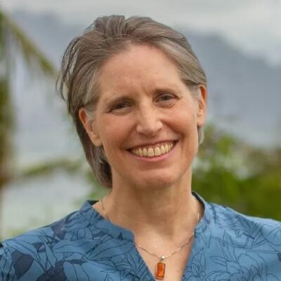 Smiling woman with short gray hair, wearing a blue patterned shirt and pendant, outdoors with greenery.