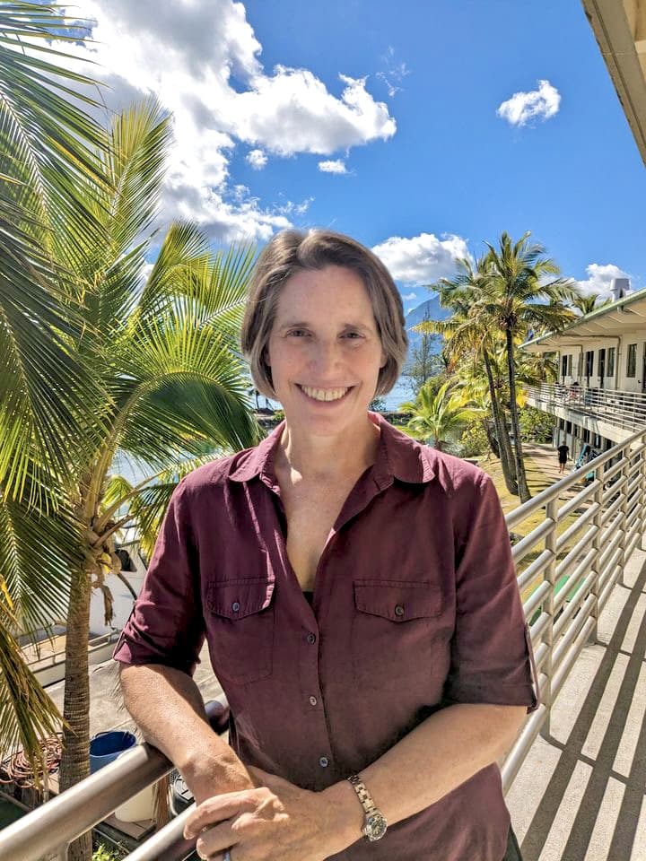 Woman in a maroon shirt smiling on a sunny balcony with palm trees and blue sky in the background.